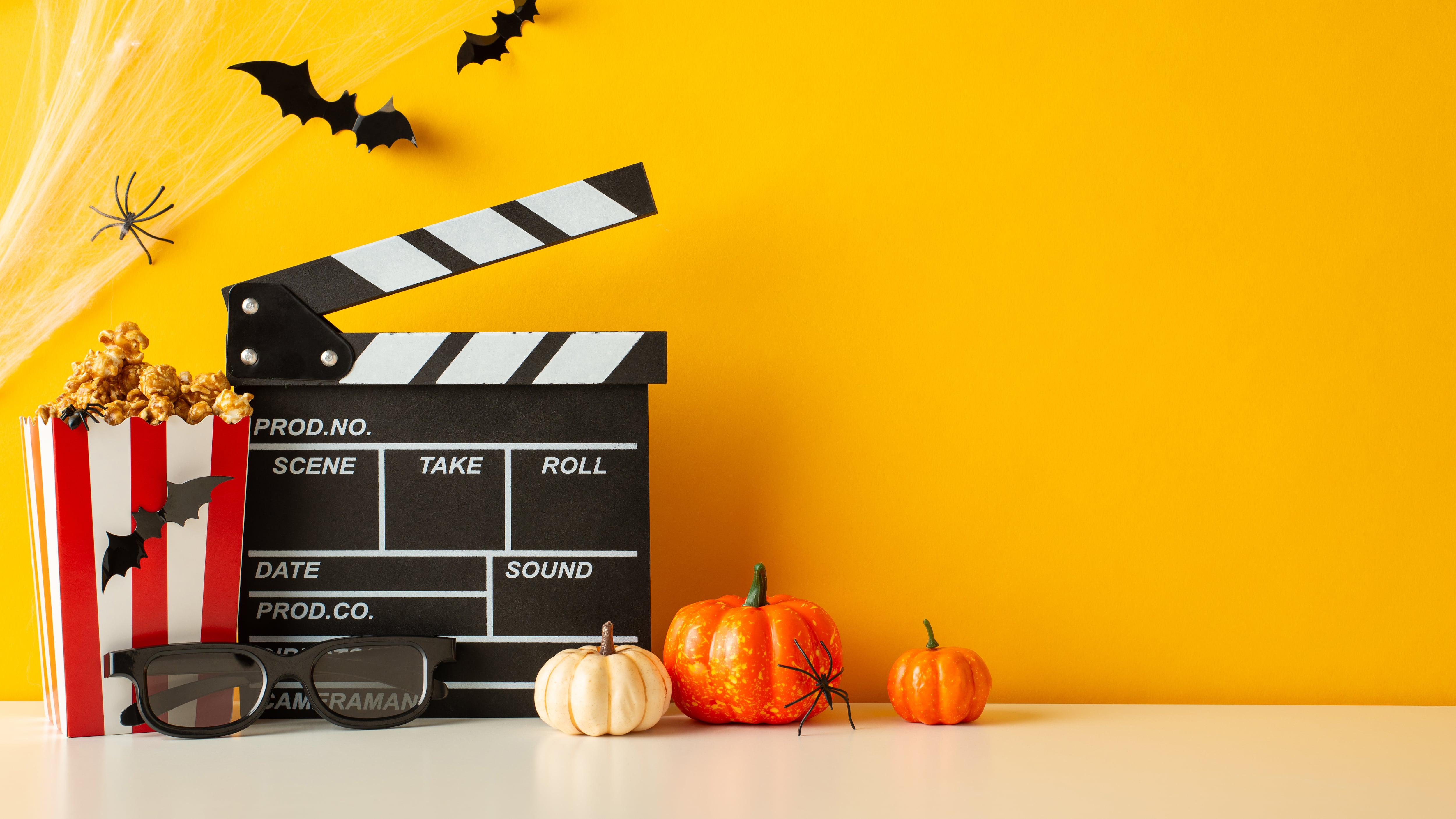 Side view shot features a table adorned with pumpkins, insects, 3D glasses, clapperboard and popcorn boxes on yellow wall background, spooky movie promo